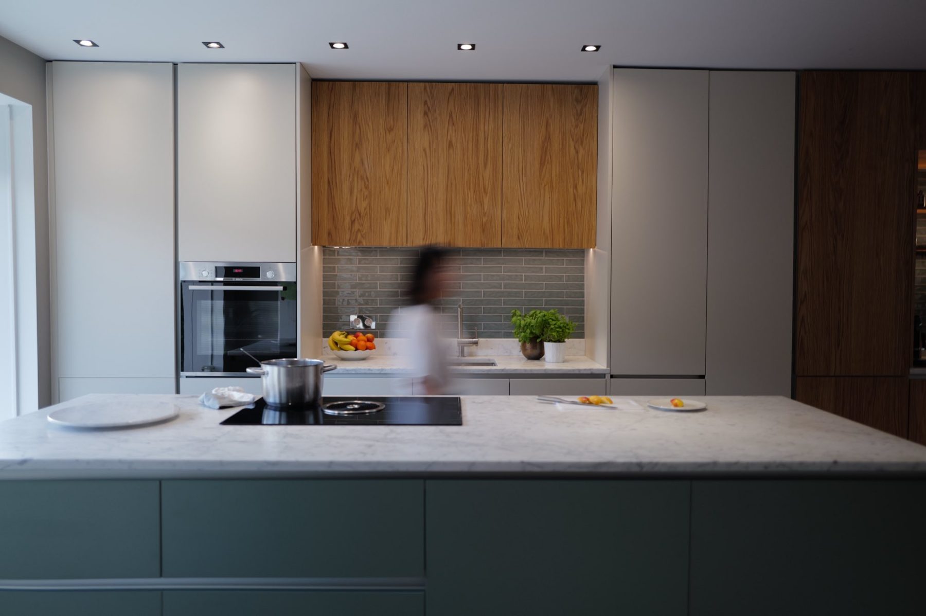 A kitchen designed by F*HD: oak upper cabinets, sage-green island with marble top, someone cooking in the background